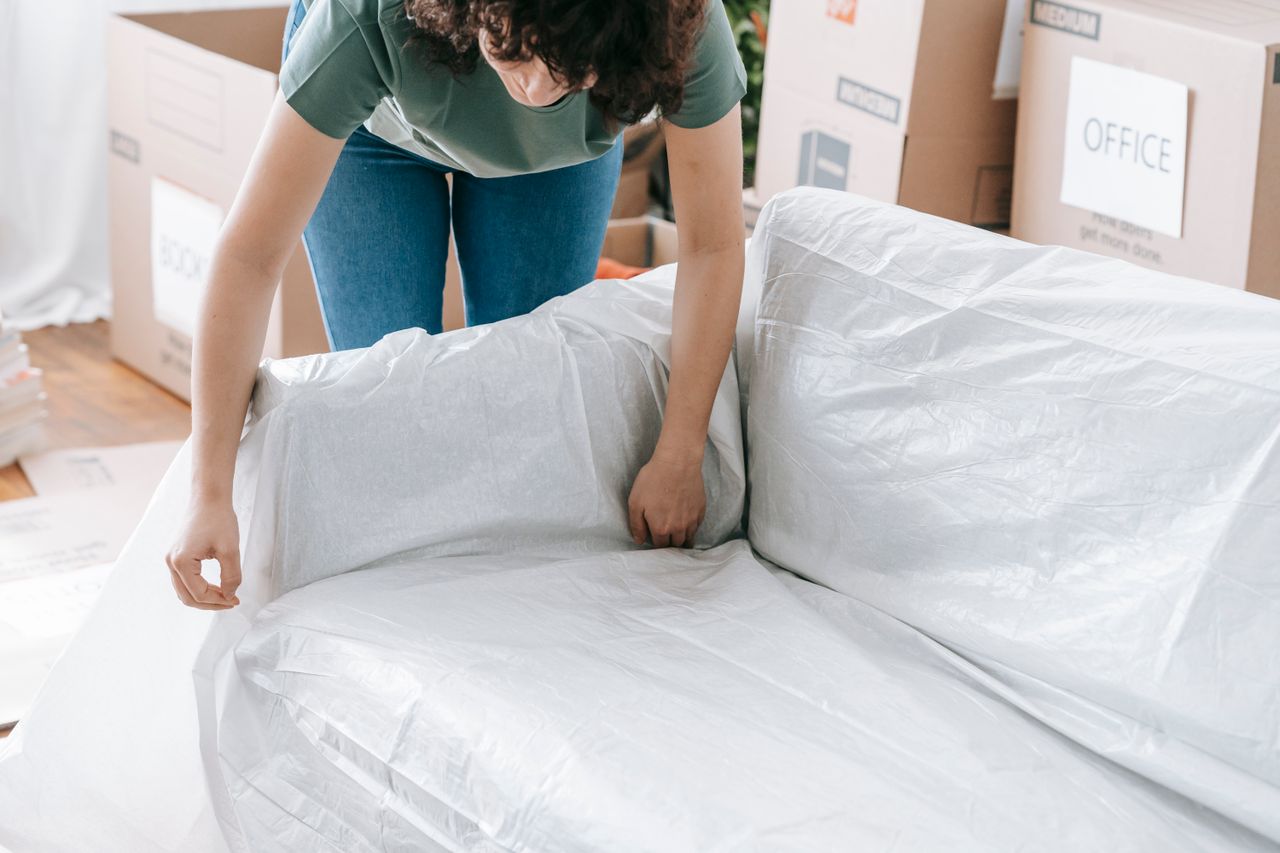 our-story A woman neatly covers a sofa with protective wrapping while boxes are being packed in a bright room.