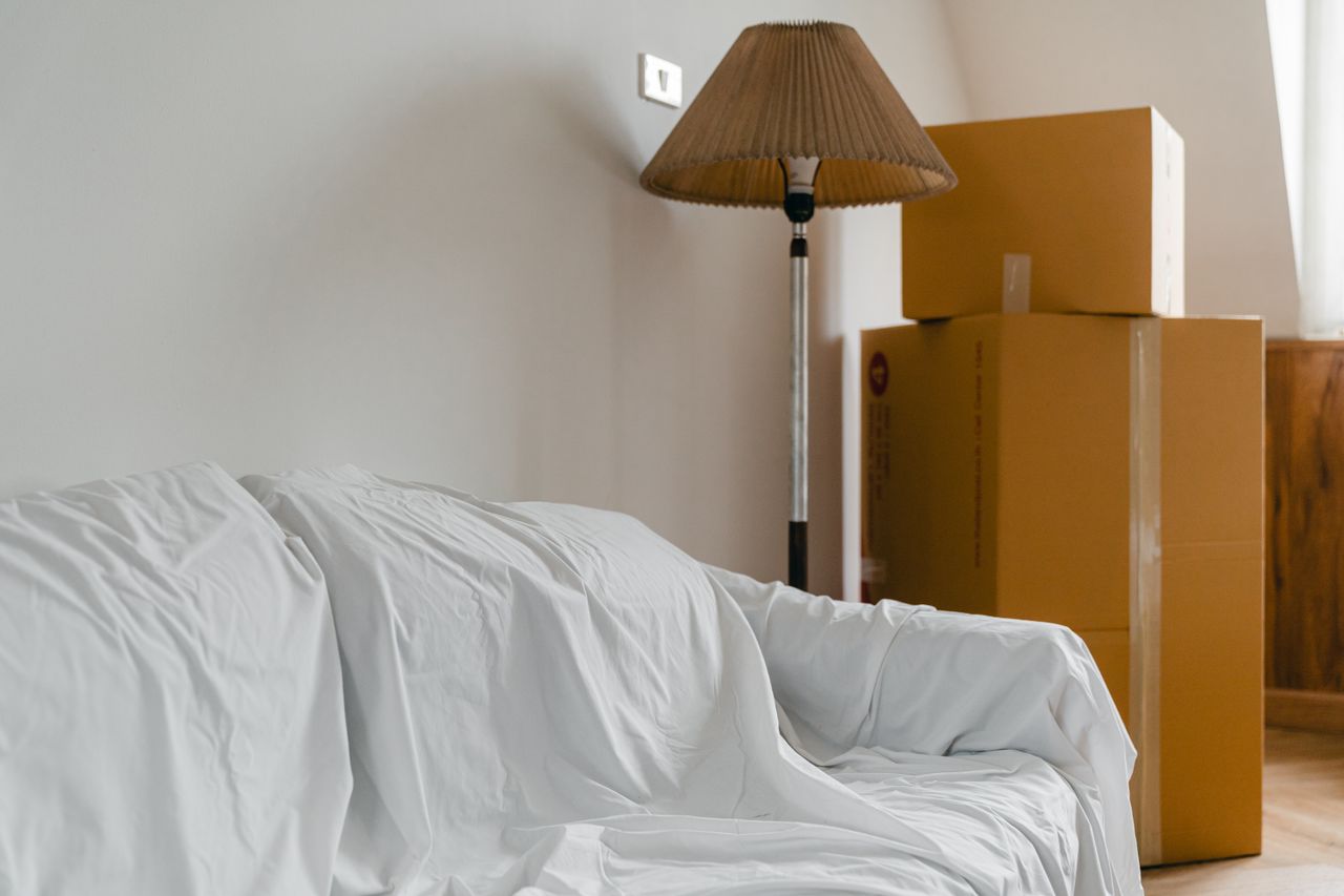 gallery-01 Canapé recouvert d’un drap, lampe sur pied et cartons emballés dans une pièce claire et spacieuse d’une maison contemporaine neuve